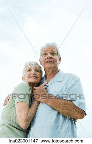 Stock Photo - Senior couple embracing and looking away, man raising eyebrows, low angle view, waist up. Fotosearch