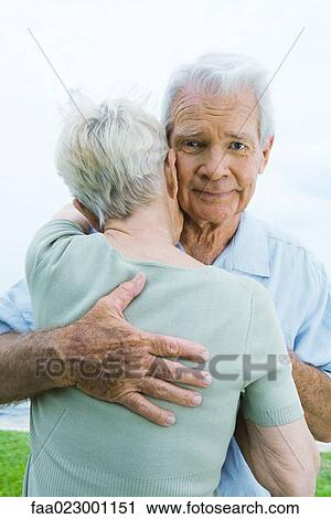 Stock Image - Senior couple hugging, man looking at camera. Fotosearch
