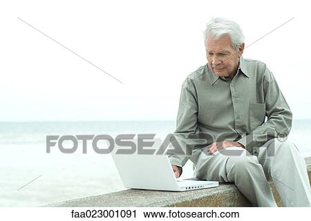 Senior man sitting outdoors using laptop computer, looking down View Large Photo Image Stock Image - Senior man sitting outdoors using laptop computer, looking down. Fotosearch