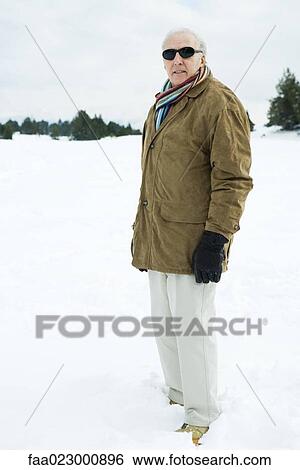 Stock Photograph - Senior man standing in snow, looking at camera, portrait. Fotosearch