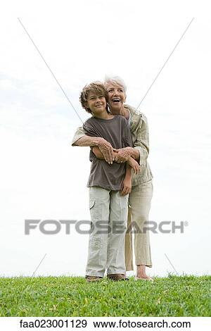 Stock Photo - Senior woman and grandson standing on grass, woman hugging boy from behind, both smiling and looking out of frame, full length. Fotosearch