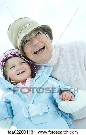 Stock Photo - Senior woman embracing granddaughter in snow, both smiling, waist up. Fotosearch