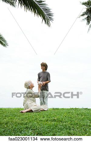 Senior woman sitting on grass, grandson handing her gift, full length View Large Photo Image Picture - Senior woman sitting on grass, grandson handing her gift, full length. Fotosearch