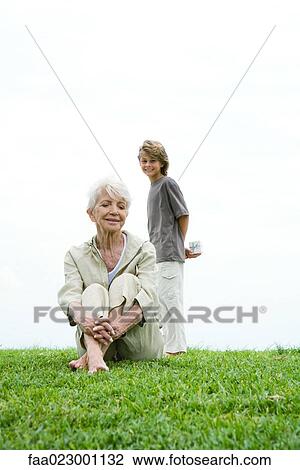 Senior woman sitting on grass, grandson standing behind her, holding gift, smiling, full length View Large Photo Image Stock Image - Senior woman sitting on grass, grandson standing behind her, holding gift, smiling, full length. Fotosearch