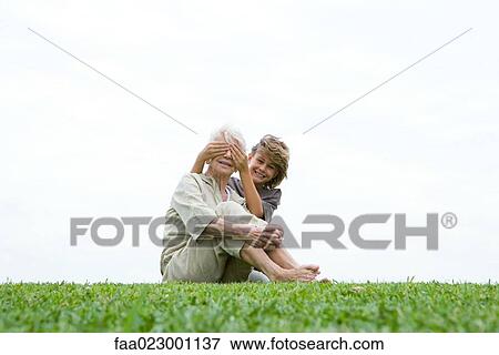 Senior woman sitting on grass, grandson covering her eyes from behind, smiling at camera, full length View Large Photo Image Stock Photo - Senior woman sitting on grass, grandson covering her eyes from behind, smiling at camera, full length. Fotosearch