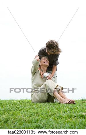 Senior woman sitting on grass, grandson leaning over her shoulder, handing her gift, full length View Large Photo Image Stock Photography - Senior woman sitting on grass, grandson leaning over her shoulder, handing her gift, full length. Fotosearch