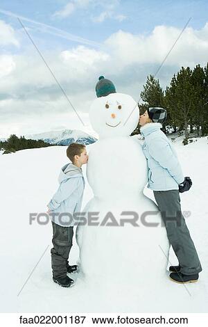 Stock Photo - Sister and brother standing, kissing snowman, side view. Fotosearch