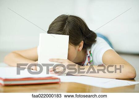 Stock Image - Teen girl lying on floor, doing homework, holding book in front of face. Fotosearch