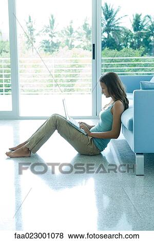Stock Photo - Teen girl sitting on the ground, leaning against sofa, using laptop computer. Fotosearch