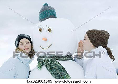 Stock Photography - Teen girls on either side of snowman, one whispering, the other listening. Fotosearch