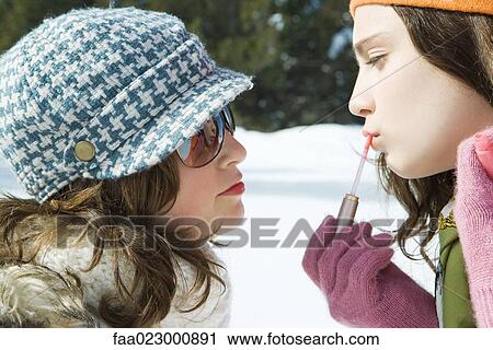 Stock Image - Teenage girl applying lip-gloss, looking at her reflection in friend's sunglasses, both smiling. Fotosearch