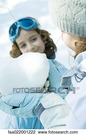 Stock Image - Teenage girl holding heart made of snow, sister smiling in background. Fotosearch