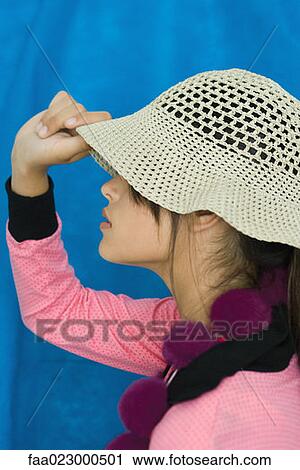 Stock Image - Teenage girl pulling sun hat over eyes, side view, portrait. Fotosearch