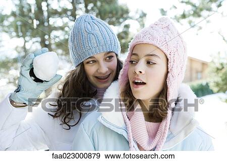 Stock Photo - Teenage girl standing behind friend, holding up snowball, smiling. Fotosearch