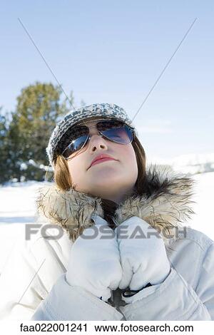 Stock Image - Teenage girl wearing parka and sunglasses in snow, head back, portrait. Fotosearch