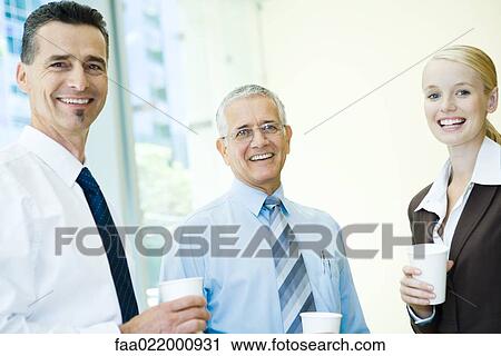 Stock Image - Three business associates smiling at camera, holding hot beverages, portrait. Fotosearch