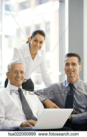 Stock Photo - Three business associates smiling at camera, group portrait, one using laptop computer. Fotosearch