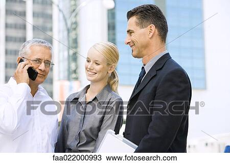 Three business associates standing together, one using cell phone, looking at camera View Large Photo Image Stock Image - Three business associates standing together, one using cell phone, looking at camera. Fotosearch