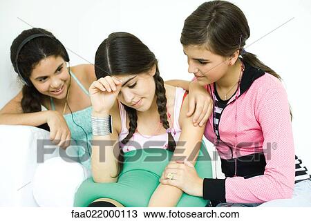 Stock Image - Three young female friends sitting on sofa, two comforting the other. Fotosearch
