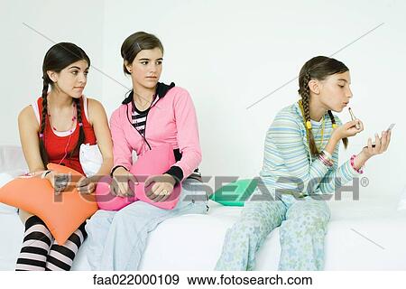Three young female friends sitting on sofa, two watching younger girl putting on make-up View Large Photo Image Stock Photo - Three young female friends sitting on sofa, two watching younger girl putting on make-up. Fotosearch