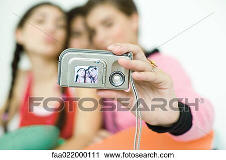 Stock Image - Three young female friends taking photo of selves with digital camera, focus on camera in foreground. Fotosearch