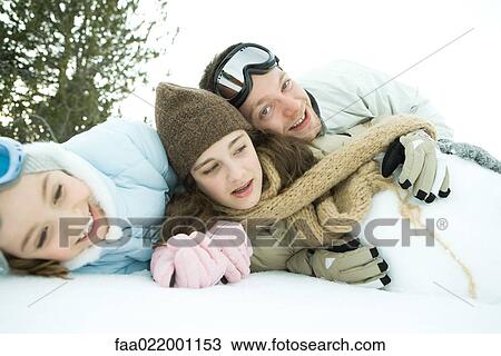 Stock Image - Three young friends reclining in snow, piled up, one smiling at camera. Fotosearch