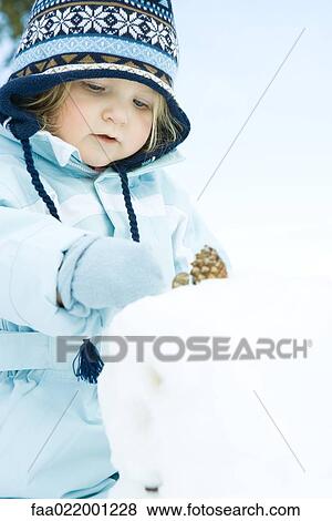 Stock Photo - Toddler girl building snowman, dressed in winter clothing. Fotosearch