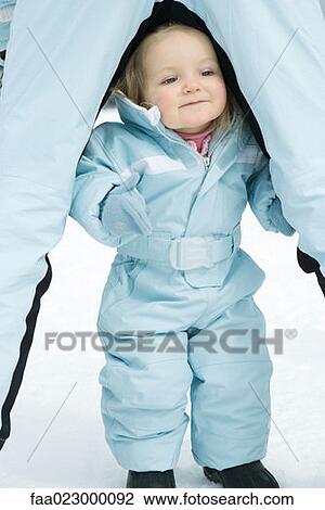 Toddler girl standing between sibling's legs, dressed in ski-suit, smiling View Large Photo Image Stock Image - Toddler girl standing between sibling's legs, dressed in ski-suit, smiling. Fotosearch