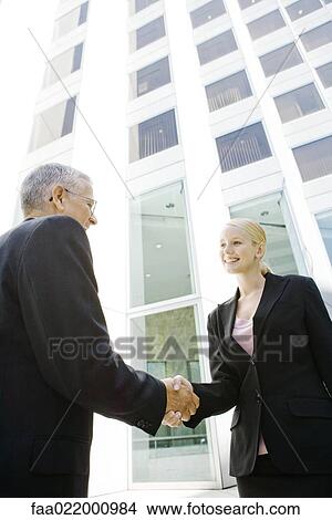 Picture - Two business associates shaking hands, smiling, building in background. Fotosearch