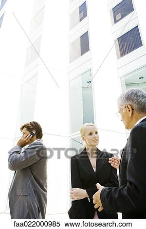 Two business associates standing, discussing, man using cell phone in background View Large Photo Image Stock Photography - Two business associates standing, discussing, man using cell phone in background. Fotosearch