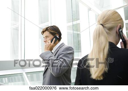 Stock Photograph - Two business associates walking past each other by window, both using cell phones. Fotosearch
