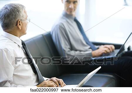 Two businessmen sitting on sofa, looking at each other, both using laptops computers View Large Photo Image Stock Photo - Two businessmen sitting on sofa, looking at each other, both using laptops computers. Fotosearch