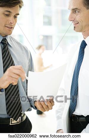 Stock Image - Two businessmen standing together, discussing document. Fotosearch