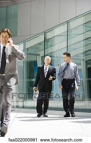 Stock Image - Two businessmen walking in front of building, discussing, one looking at camera. Fotosearch