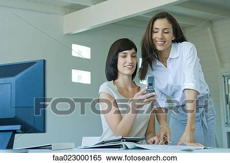 Stock Photography - Two female co-workers, one showing the other cell phone. Fotosearch