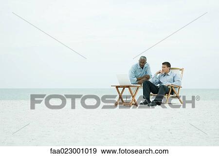 Two men on the beach, looking at laptop computer together, full length View Large Photo Image Stock Photo - Two men on the beach, looking at laptop computer together, full length. Fotosearch