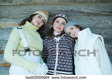 Picture - Two sisters and a friend dressed in winter clothes, leaning against log wall, portrait. Fotosearch