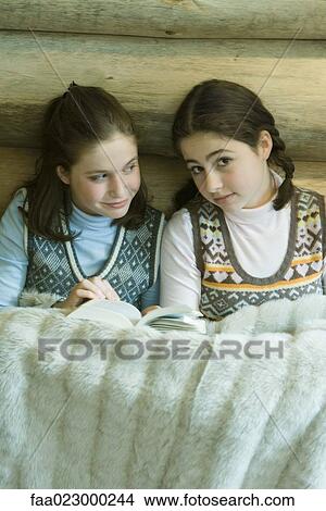 Picture - Two teen girls in winter clothes sitting under warm blanket together. Fotosearch