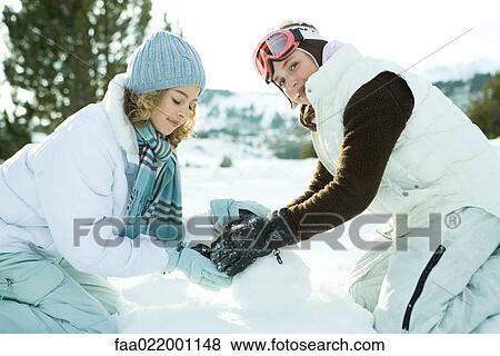 Stock Photo - Two teenage girls crouching in snow, making snowball together, one smiling at camera. Fotosearch