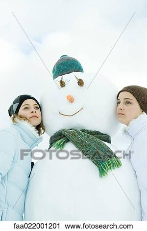 Stock Image - Two teenage girls leaning against snowman, looking away, portrait. Fotosearch