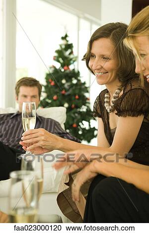 Stock Image - Two women and man sitting on couches, drinking champagne, Christmas tree in background. Fotosearch