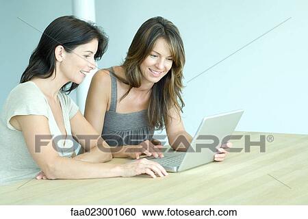 Two women sitting at table, looking at laptop computer together, waist up View Large Photo Image Stock Image - Two women sitting at table, looking at laptop computer together, waist up. Fotosearch