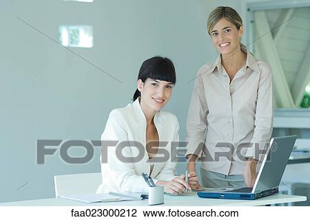 Two young businesswomen in office, one seated at desk, the other standing by her side, both smiling at camera, portrait View Large Photo Image Stock Image - Two young businesswomen in office, one seated at desk, the other standing by her side, both smiling at camera, portrait. Fotosearch