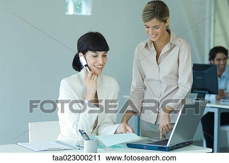Two young businesswomen, one sitting at desk, using cell phone, the other standing next to her, touching laptop, both smiling View Large Photo Image Stock Image - Two young businesswomen, one sitting at desk, using cell phone, the other standing next to her, touching laptop, both smiling. Fotosearch