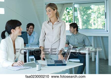 Stock Image - Two young businesswomen smiling at each other in office, male colleagues in background. Fotosearch