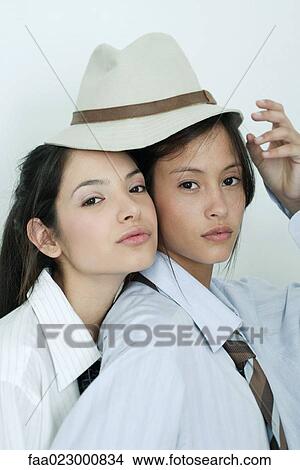 Picture - Two young female friends dressed in button down shirts, ties, one resting head on the other's shoulder, sharing hat, portrait. Fotosearch