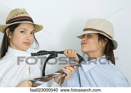 Stock Image - Two young female friends dressed in hats, pulling each other's ties, one frowning, the other smirking. Fotosearch