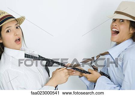 Stock Image - Two young female friends dressed in hats, pulling each other's ties, looking at camera. Fotosearch