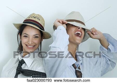 Stock Image - Two young female friends dressed in button down shirts, ties and hats, laughing, portrait. Fotosearch