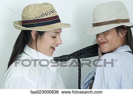 Stock Photograph - Two young female friends dressed in button down shirts, ties and hats, one pulling on the other's tie, portrait. Fotosearch